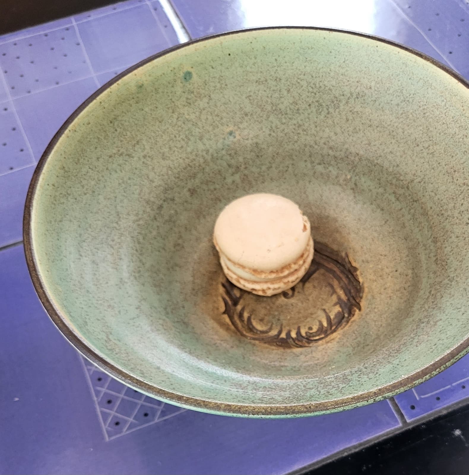 A teal-green stoneware bowl with an impressed chop-mark seal at its center, holding a single macaron.