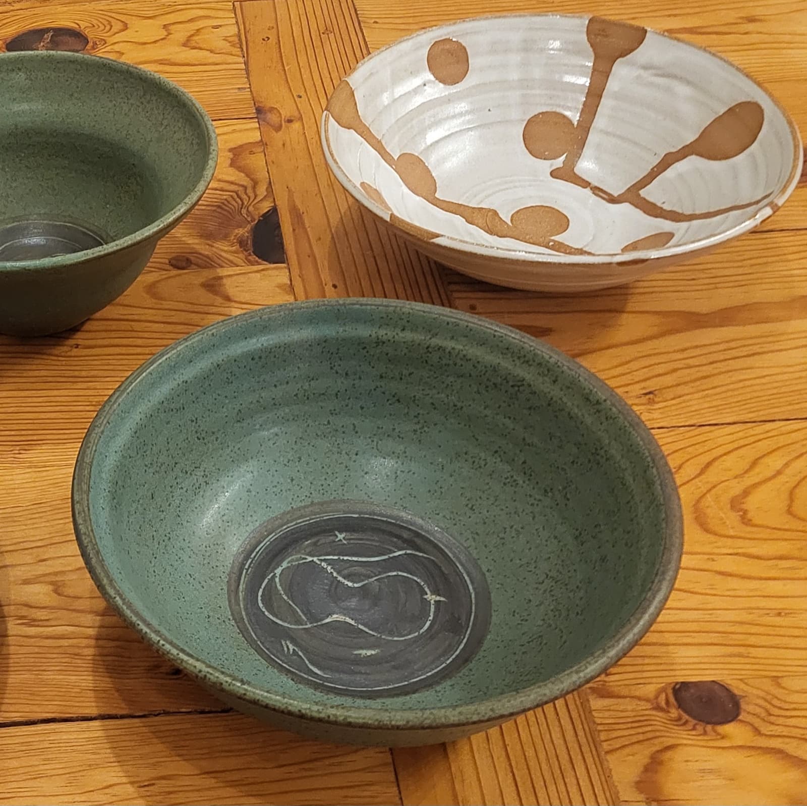 Three stoneware bowls arranged on a wood floor: two green, one ivory with rust-brown lollipop motifs.