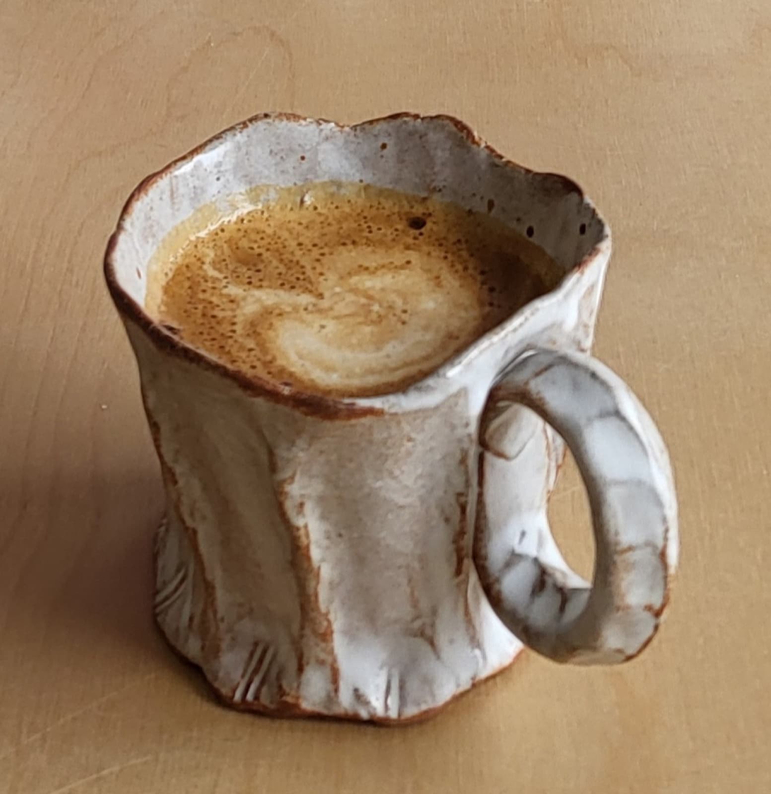 A pinched stoneware coffee mug holding espresso, with a cream and warm-earth glaze on a wood surface.